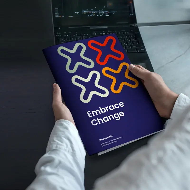 Hands holding finished full-colour booklet with Embrace Change cover beside laptop; strong finishing/application image.